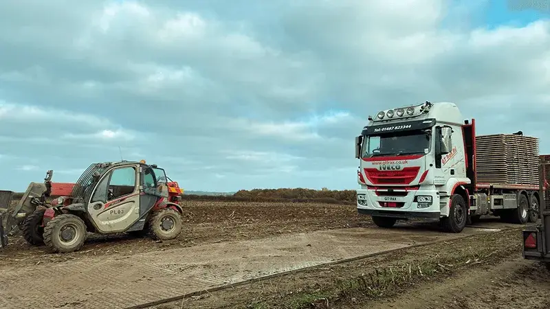 Red telehandler on a muddy TuffTrak track next to a white flatbed truck loaded with more TuffTrak panels in a field under a cloudy sky.