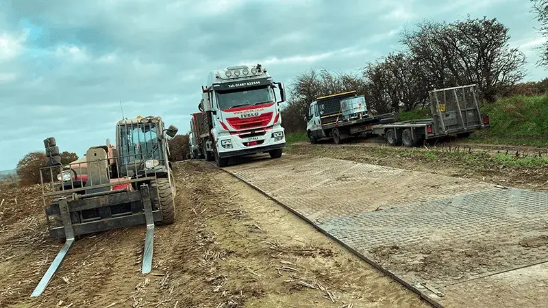 Forklift and large white truck on muddy access road. Truck drives on TuffTrak panels. Flatbed truck with trailers in background. Overcast sky.