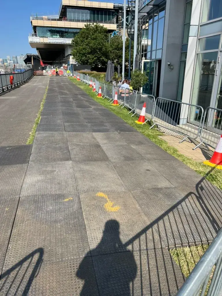 Temporary path of dark WalkOver mats with yellow marks, lined by barriers & cones, leading past an outdoor cafe and waterfront construction.