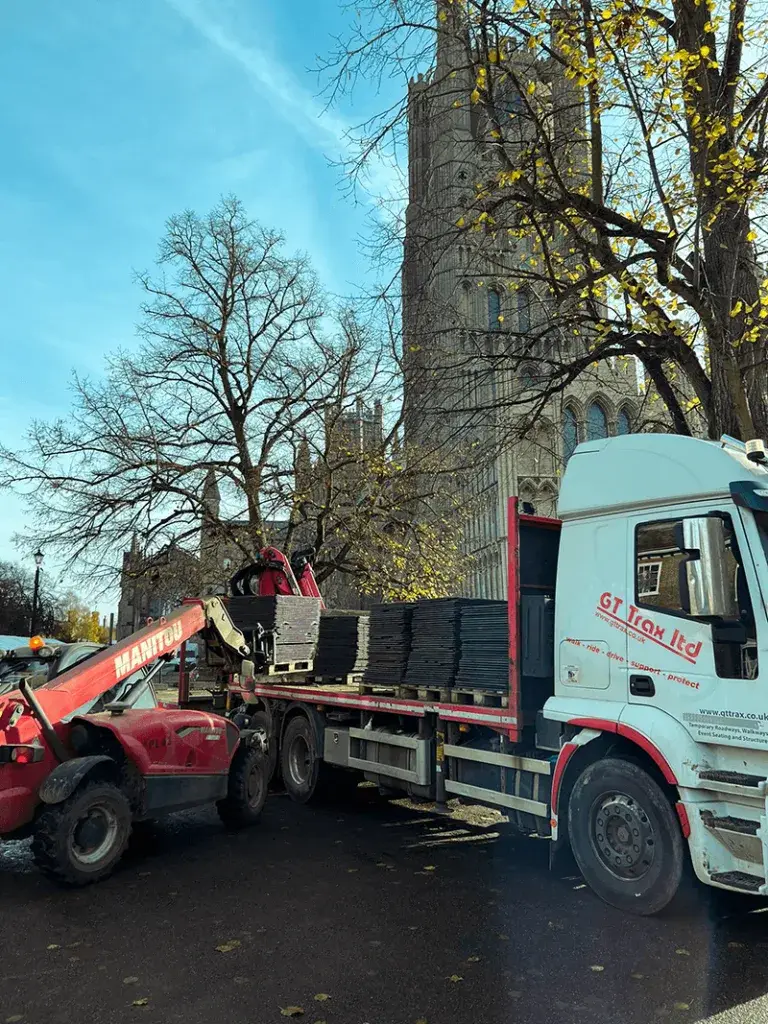 Red Manitou telehandler loading WalkOver ground protection mats onto a white GT Trax flatbed truck near a stone cathedral and bare trees.