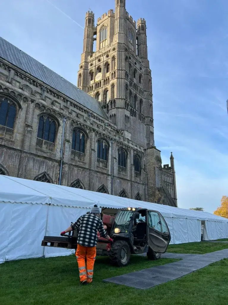 Man in orange pants guides a small forklift carrying WalkOver ground mats next to a long white tent and a large historic cathedral.