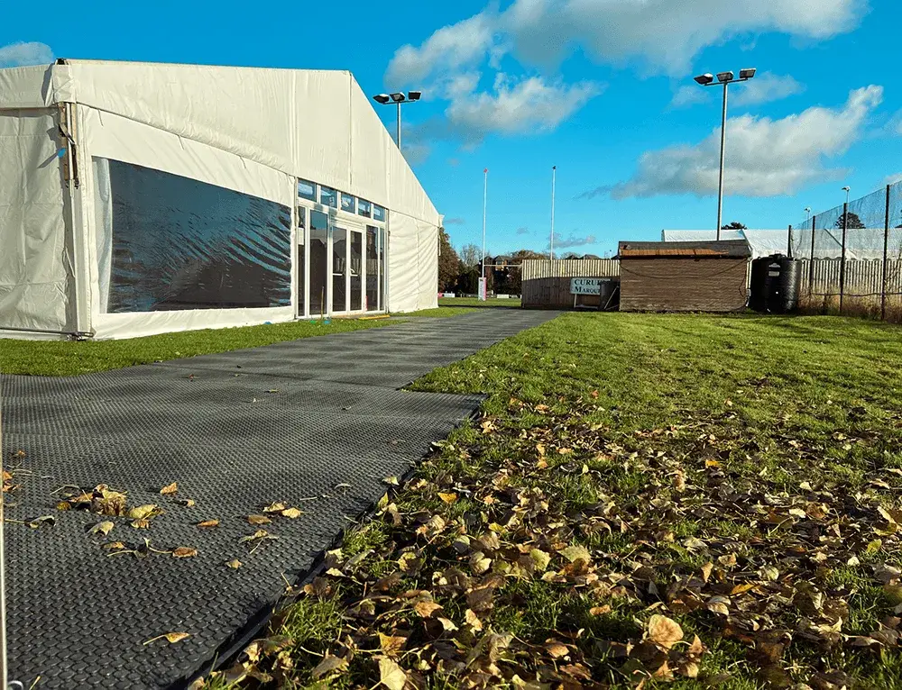 A white event tent with glass doors and a WalkOver path on a grassy field with autumn leaves. Floodlights and a shed are in the background.