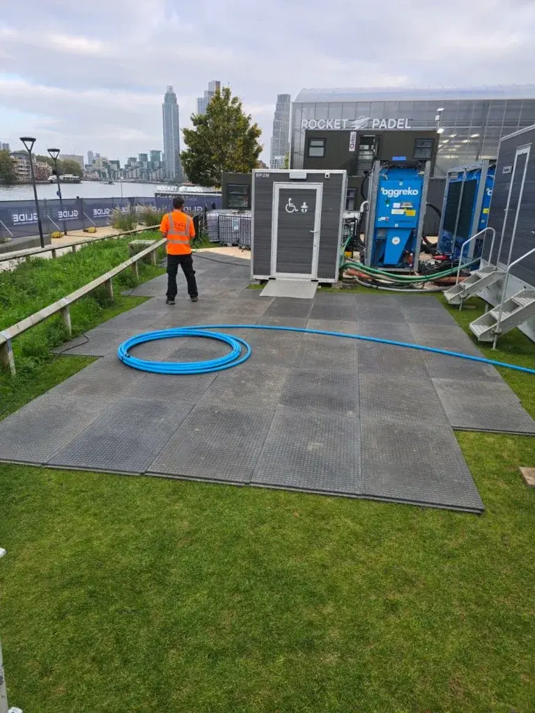 Person in orange vest near portable accessible restroom, blue machinery, and hoses on outdoor WalkOver mats. River and city skyline.