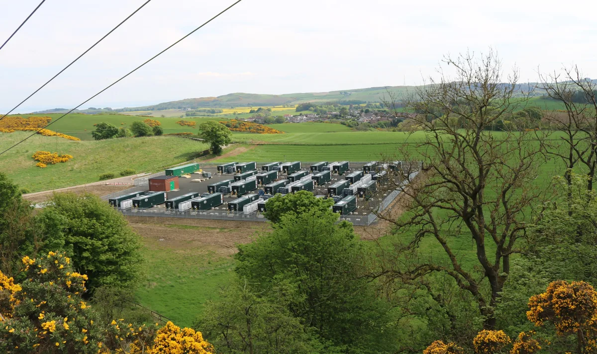 The Camilla battery energy storage facility with rows of white container units, gravel access roads, and electrical infrastructure in a rural UK setting.