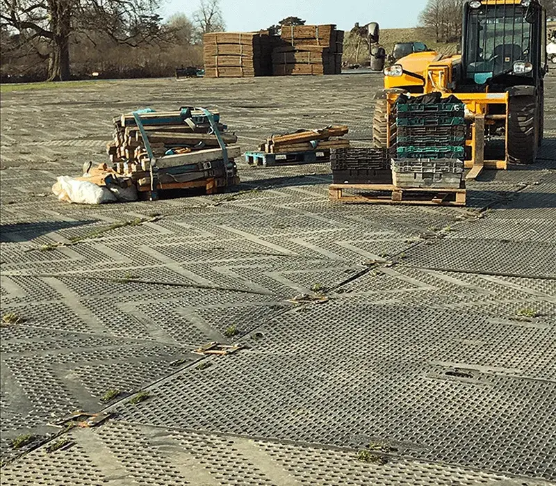 Dark grey perforated EuroMats ground mats cover an outdoor area, with stacks of wood, crates, and a yellow forklift.
