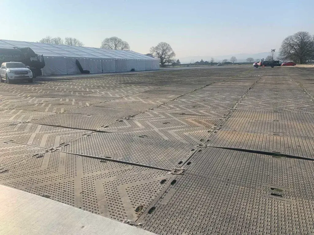 Grey interlocking EuroMats ground protection mats cover a wide field. A long white tent and vehicles in the background.