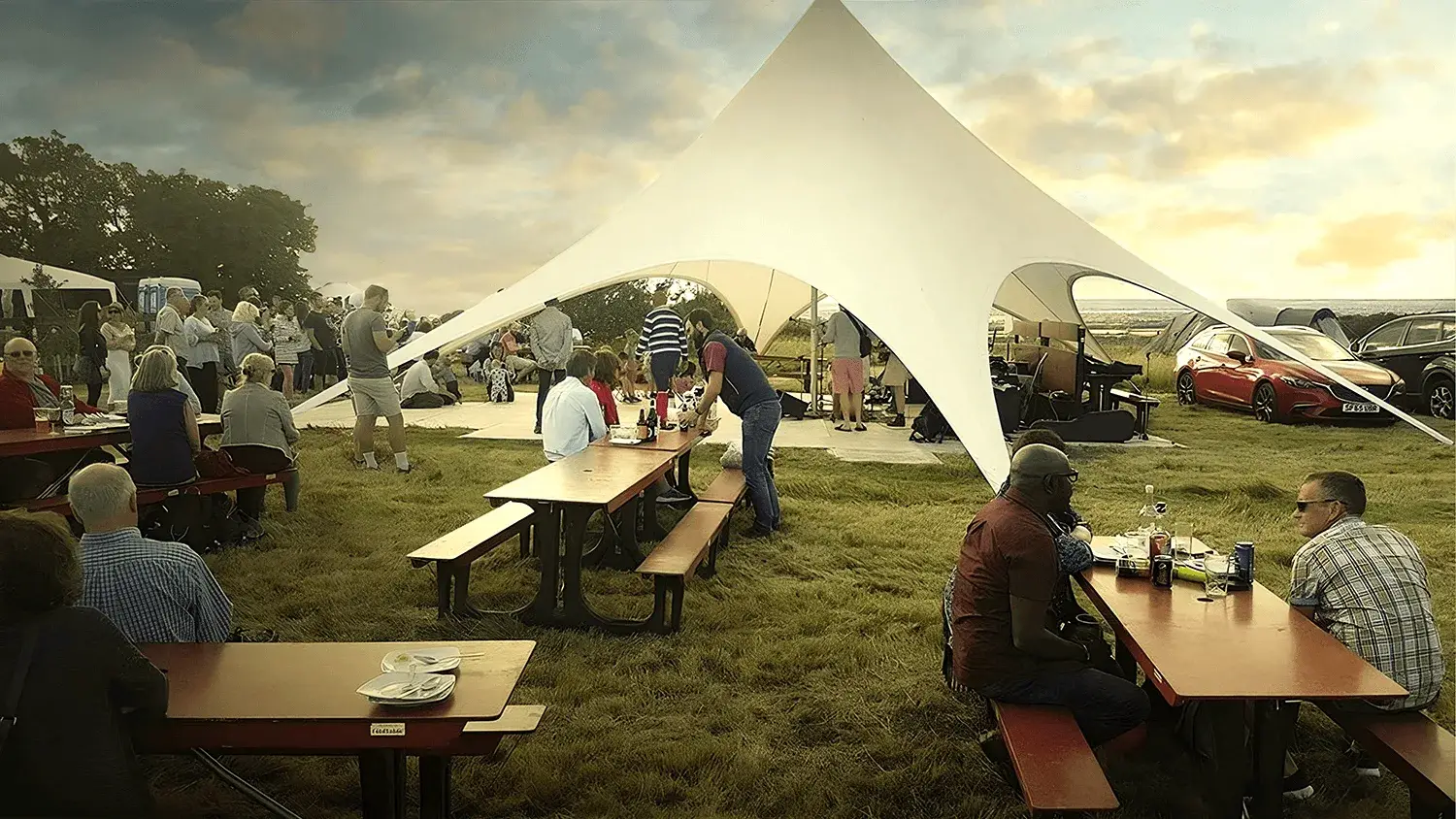 People gather under a white StarShade tent and around FoldTable picnic tables in a grassy field. Cars are visible in the background.