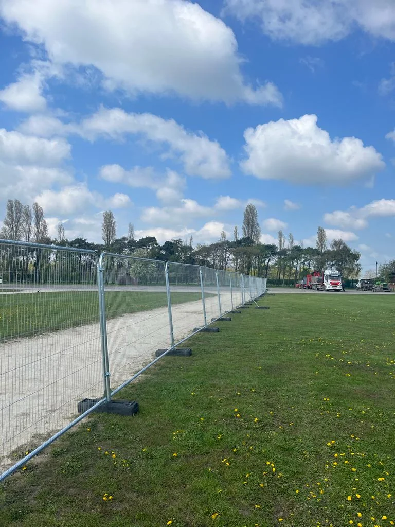 Temporary wire Heras fencing separates a grassy field from a dirt path. Cloudy blue sky, trees, and a white truck in background.