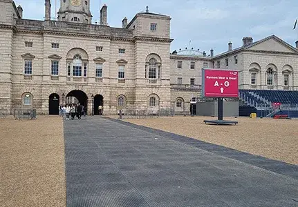 Grand stone building with people exiting archway onto sandy ground with grey EuroMats path. Red digital sign indicates "Partners Meet & Greet A-G".