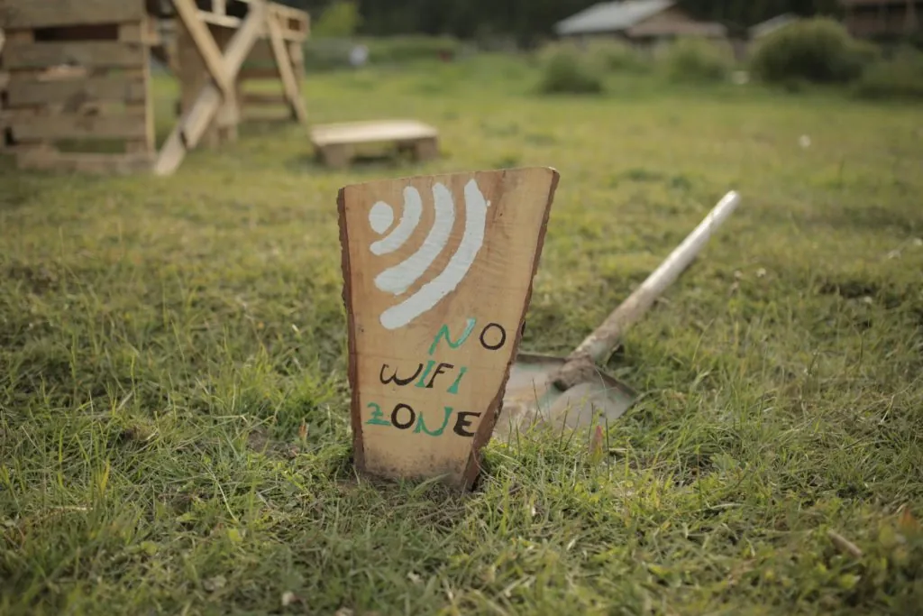 Rustic wooden "No Wifi Zone" sign with a WiFi symbol, stuck in a grassy field next to a shovel.