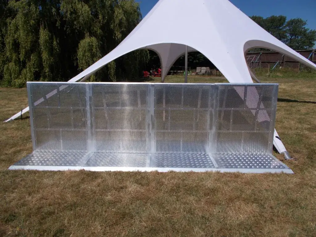 Three-section metal Stage & Pit barrier with perforated panels and textured base on dry grass. White star-shaped tent behind.