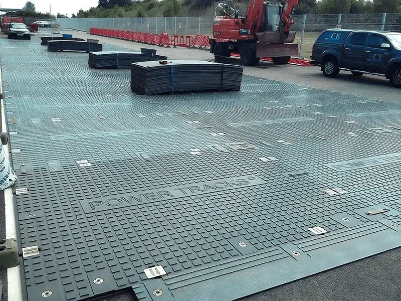 Dark grey interlocking PowerTrax 40 ground protection mats cover a large area. Stacks of mats, a red excavator, and a blue pickup truck are in the background.