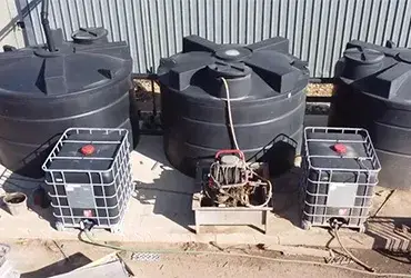 Three large black water tanks, two smaller tanks in cages, and a central pump setup outdoors by a metal fence.