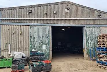 Weathered corrugated metal storage building with large open doors. Crates of produce and wooden pallets outside.