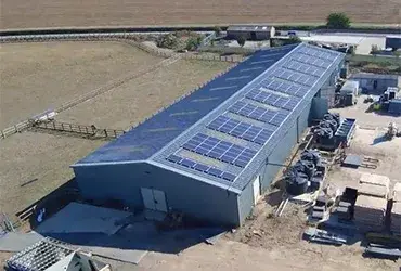 Aerial view of a long grey building with solar panels covering its roof, bordered by a fenced field and a works yard.