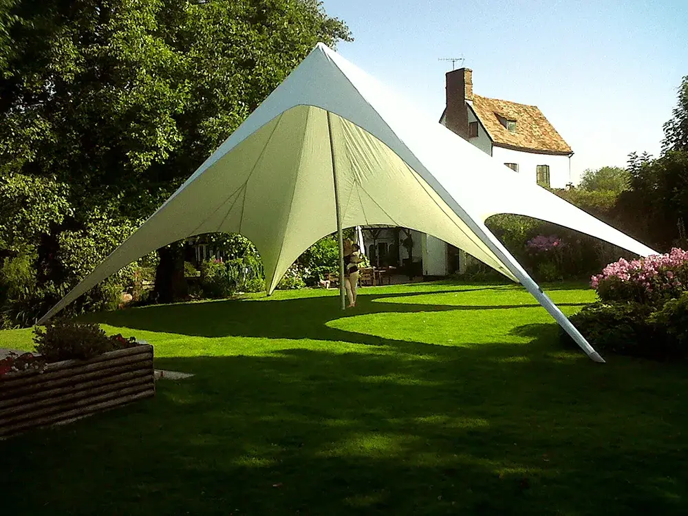 White multi-pointed StarShade tent on a sunny green lawn. A person stands under it. A house and trees are in the background.