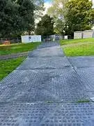 A long, textured, dark gray TuffTrak pathway extends through a grassy park with white buildings and trees in the distance.