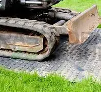 Close-up of mini-excavator track and blade resting on a black textured TuffTrak ground protection mat on green grass.