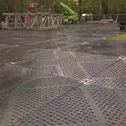 Dark interlocking TuffTrak temporary ground protection mats cover an outdoor area with a green crane and structures in the background.