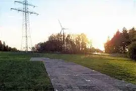 Electricity pylon and TuffTrak path in grassy field with wind turbine and trees under bright sun.