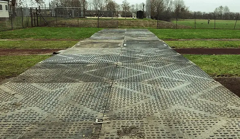 Long path of interlocking textured dark grey TuffTrak mats on a green field with a dirt track and fences.