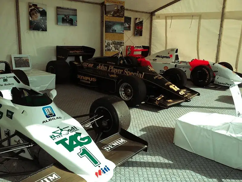 Three F1 race cars: black Lotus, white Williams, and white McLaren, on display in a tented exhibition space, on LiteTrax GridMat.