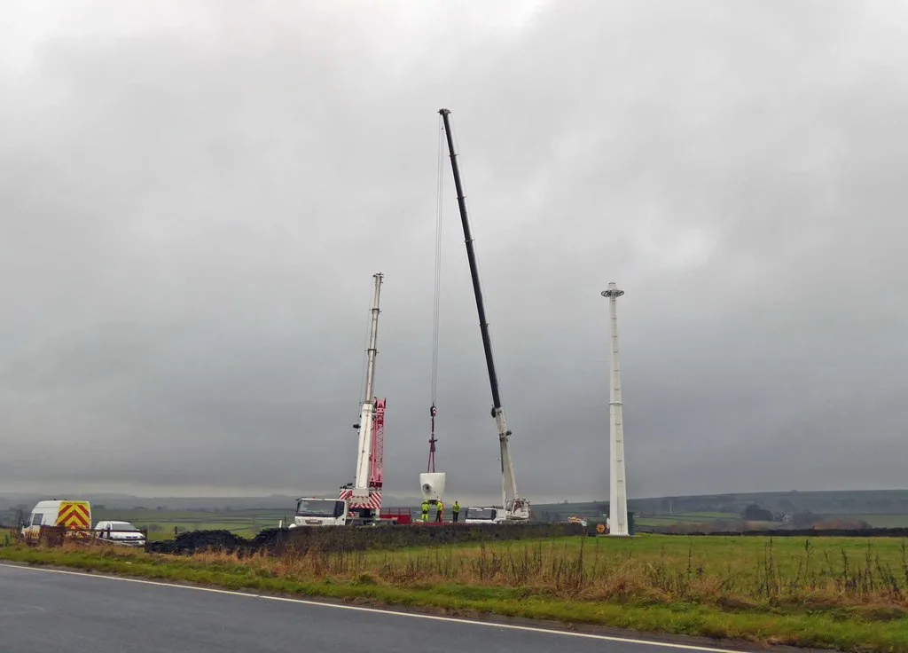 Wind turbine under construction at a UK site with crane and heavy machinery on prepared hardstanding, set against rolling countryside.