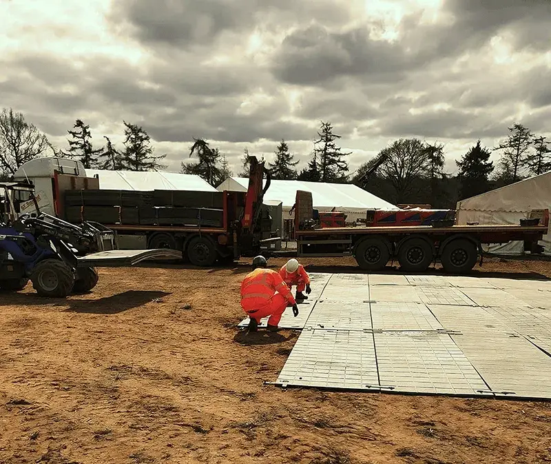 Two construction workers in orange jumpsuits lay modular PowerTrax 20 flooring on a dirt field. Flatbed trucks, a loader, and white tents behind.