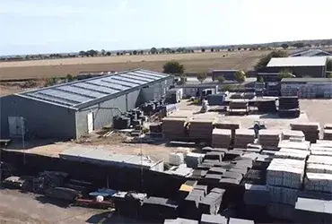 Large grey warehouse alongside an outdoor yard packed with stacks of paving stones and building materials. Rural setting with fields.